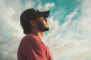 A young man in a red t-shirt and blue baseball cap staring upward with a cloud background.