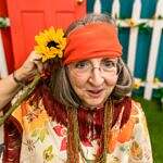 Image of an older woman with glasses holding a sunflower to her hair dressed as a "hippy".