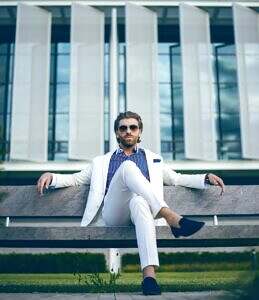 Man dressed in a white suit and sun glasses sitting on a bench confidently in front of an office building.