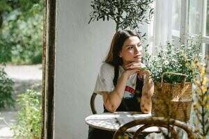 Woman sitting at a table surrounded by plants looking contemplative.