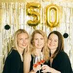 Redefining Beauty and Value After 50. Image of three women standing in front of a 50 balloon with champagne toasting.