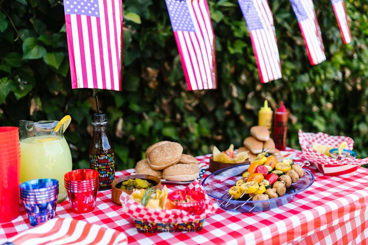 So this Memorial Day, as you dig into your favorite dishes and dodge those family debates, remember: the real flavor comes from kindness. Image of a picnic table full of food and drink with American flags hanging above.
