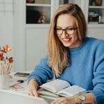 Image of a woman wearing a blue sweater sitting at a desk with book open in front of her and she is writing on a computer.