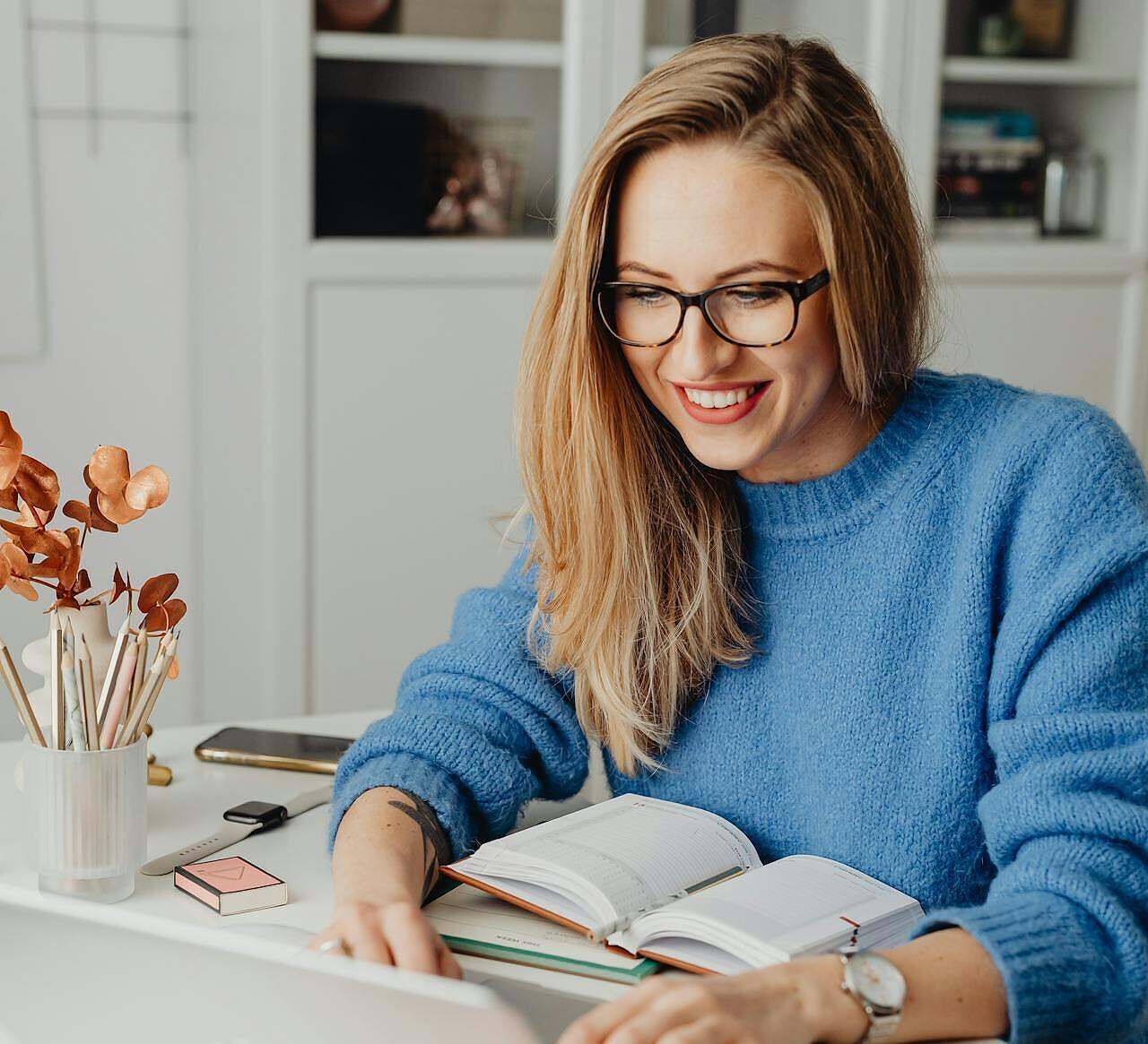 Image of a woman wearing a blue sweater sitting at a desk with book open in front of her and she is writing on a computer.