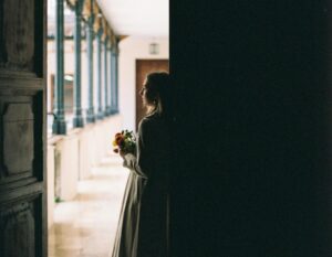 Image of a woman standing in a darkened threashold looking towards a lighted window.