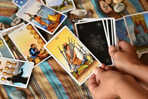 Image of tarot cards spread over a table with the five of swords being held by hands with the remaining deck unseen.