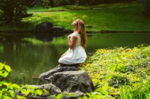 Image of a woman kneeling in prayer by the side of water looking upward towards the sun.