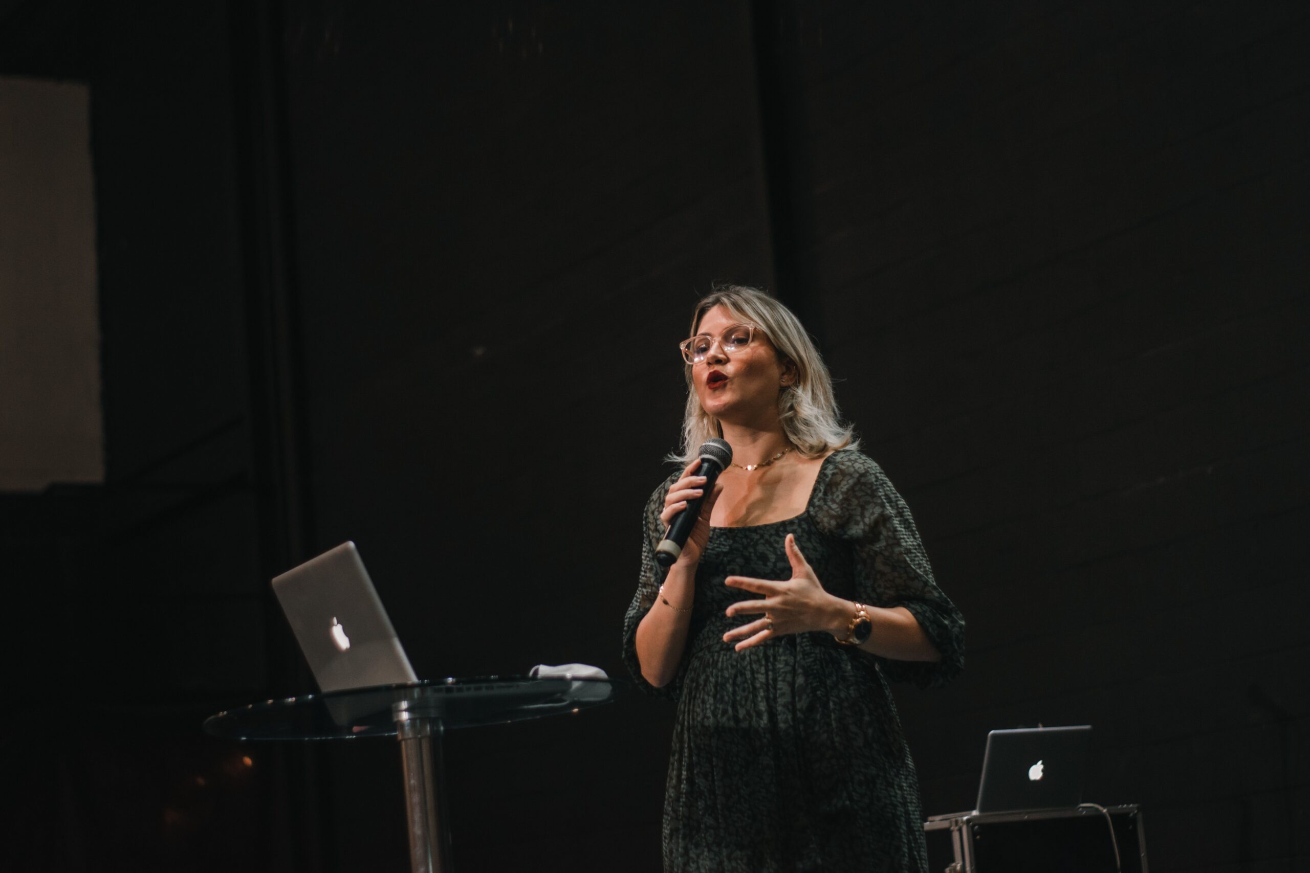 Image of a woman standing on a platform with a microphone speaking to an audience.