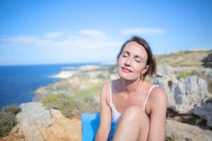 Image of a woman sitting in the sun high on a cliff overlooking the ocean.
